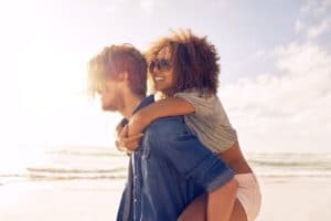 Side view portrait of young man carrying his girlfriend on his back at the beach. Boyfriends giving piggyback ride to his beautiful girlfriend at seashore.